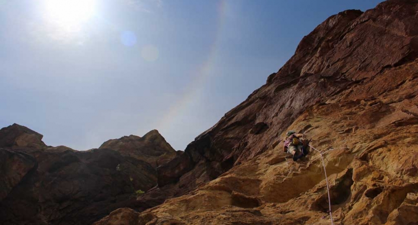 Wearing safety gear and secured by ropes, a person climbs a steep rock wall. There is a clear blue sky above them. 